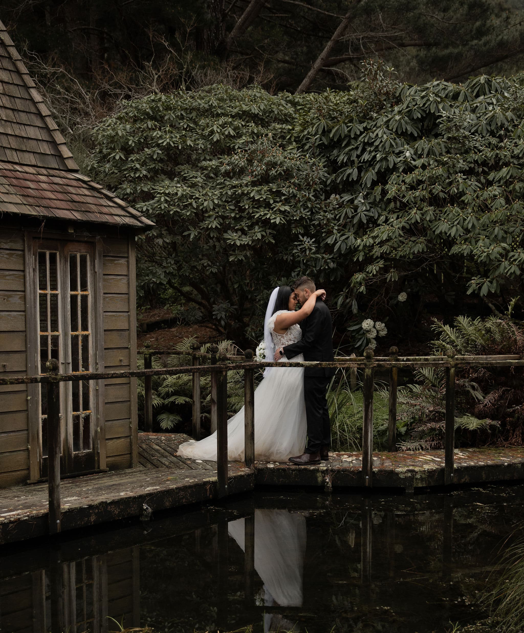 Bride and groom kiss by a rustic wooden cabin next to a pond and lush greenery.