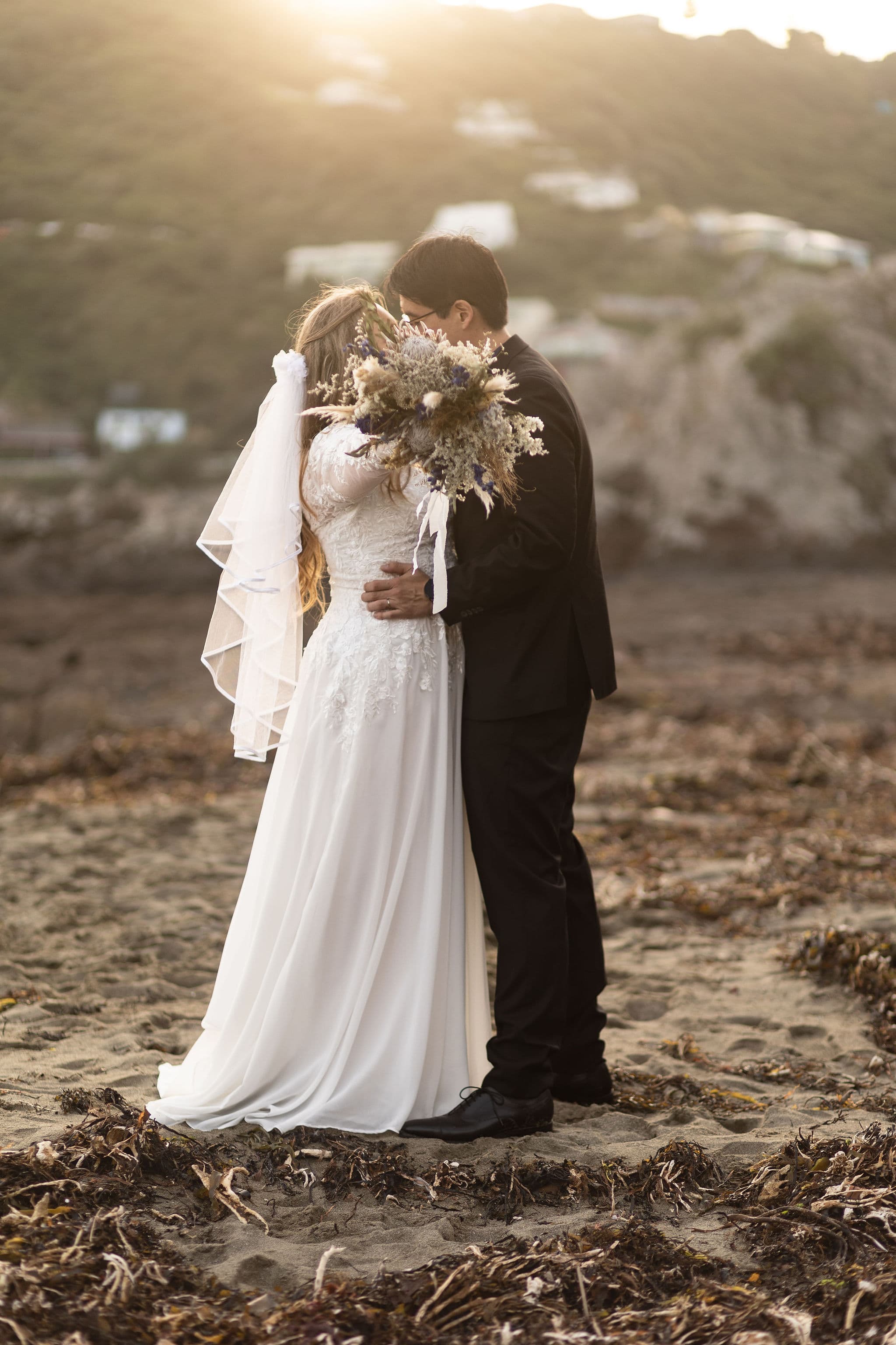 A bride in a white gown and a groom in a black suit embrace on a beach at sunset, with the bride holding a bouquet that obscures their faces and a rocky, coastal landscape in the background.