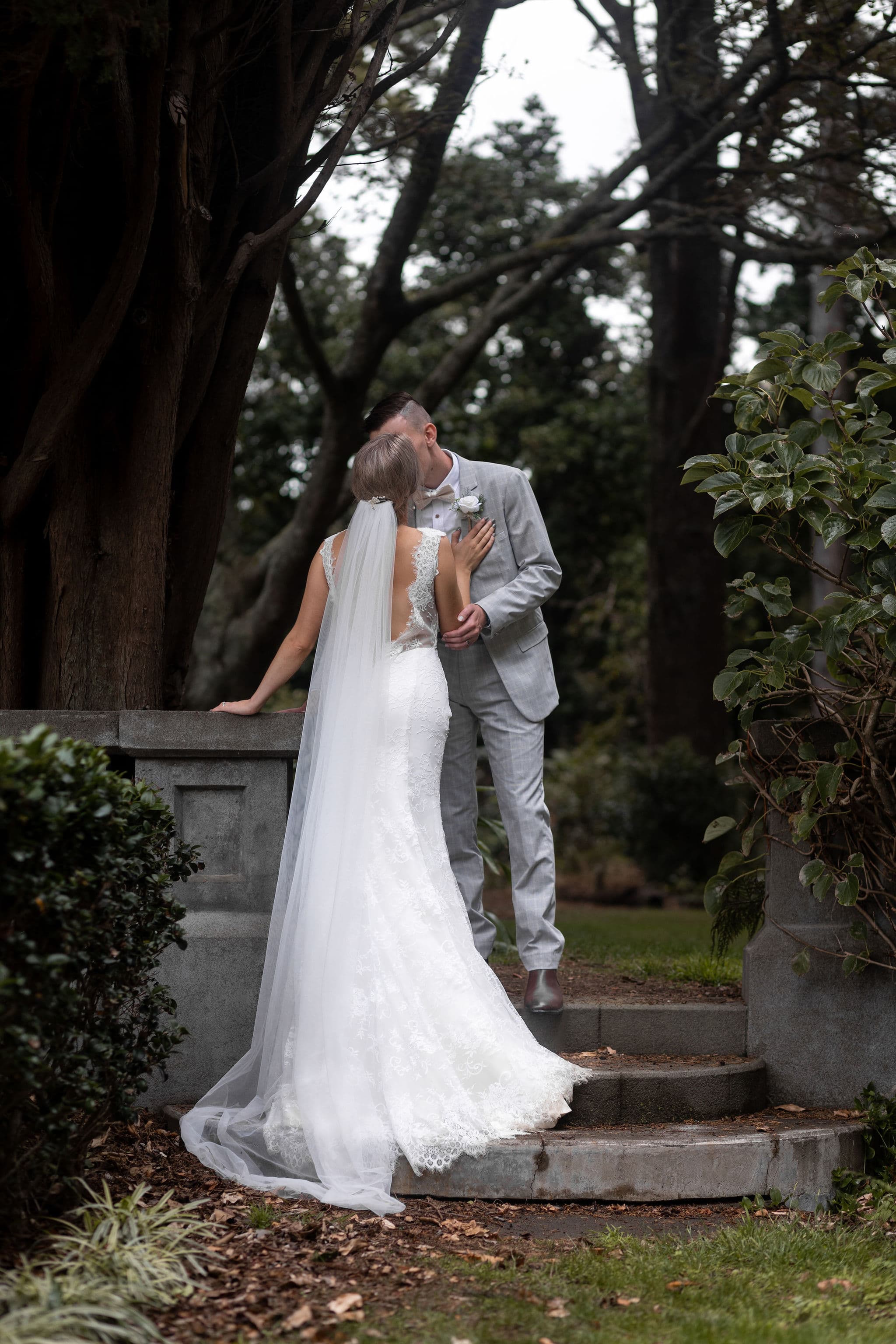 A bride and groom share a kiss outdoors on stone steps surrounded by trees and greenery.